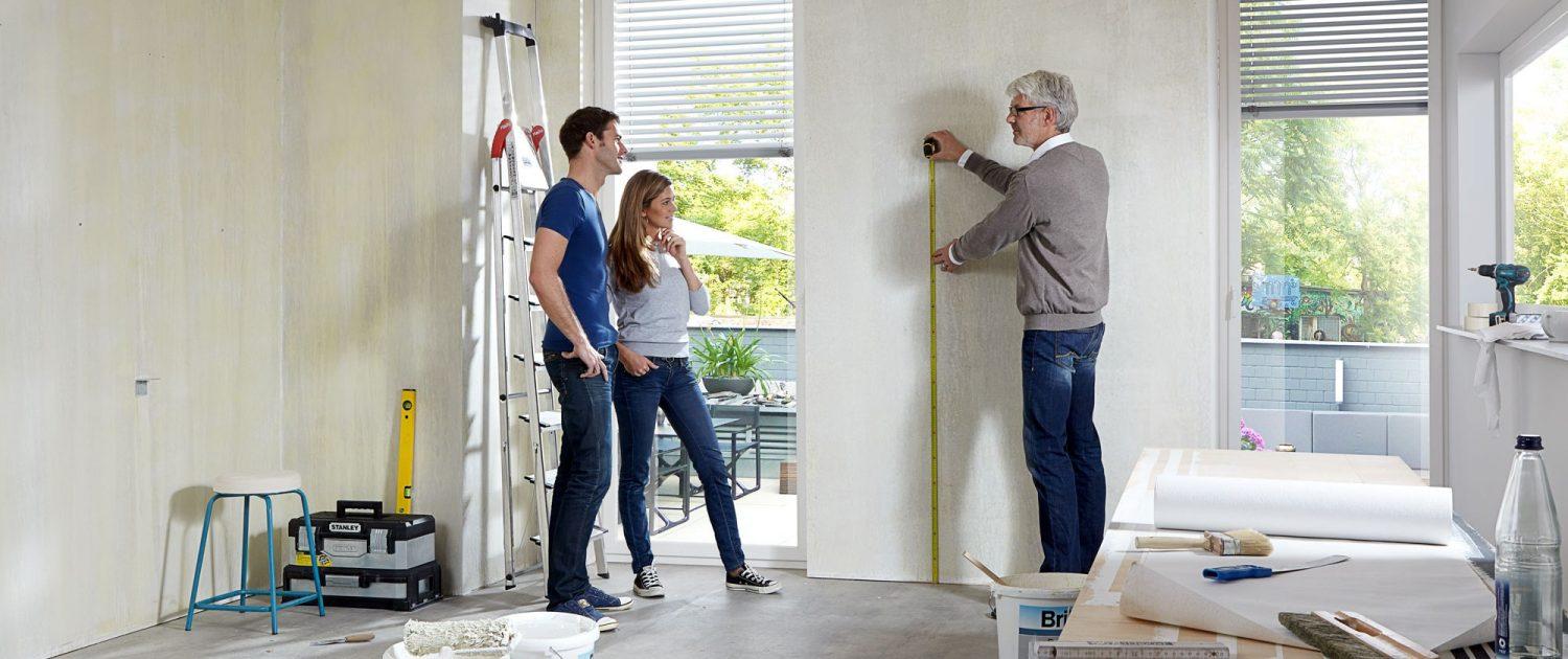 Ein Mann misst eine Wand aus und ein anderer Mann und seine Frau schauen dabei zu.