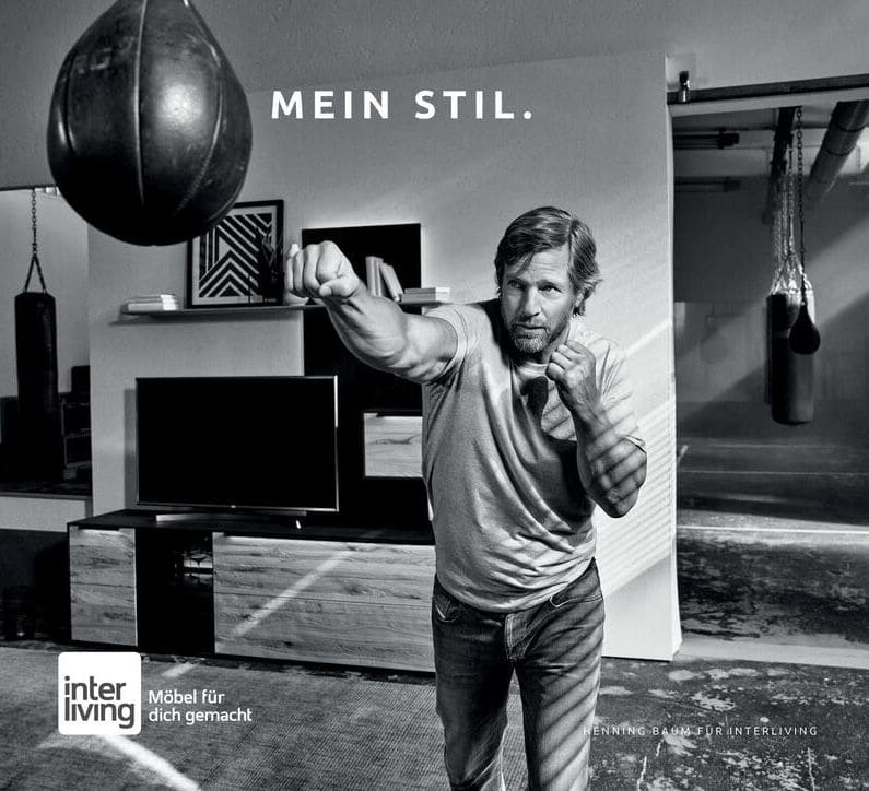 Schwarz-Weiß Bild eines Mannes, der gegen einen Boxsack boxt, hinter ihm eine moderne Wohnwand